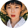Woman with short hair and freckles wearing a large sun hat, surrounded by green foliage, conveying a sense of travel and adventure related to passport services.